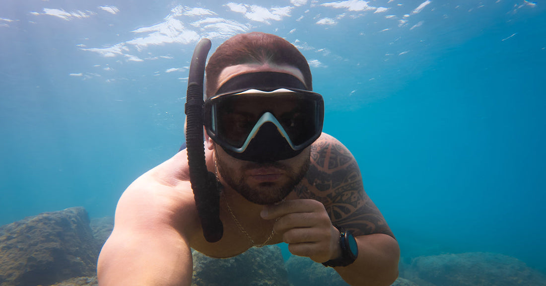 Close-up of a snorkeler underwater with mask and snorkel, arm extended toward the camera, rocky ocean floor visible below.
