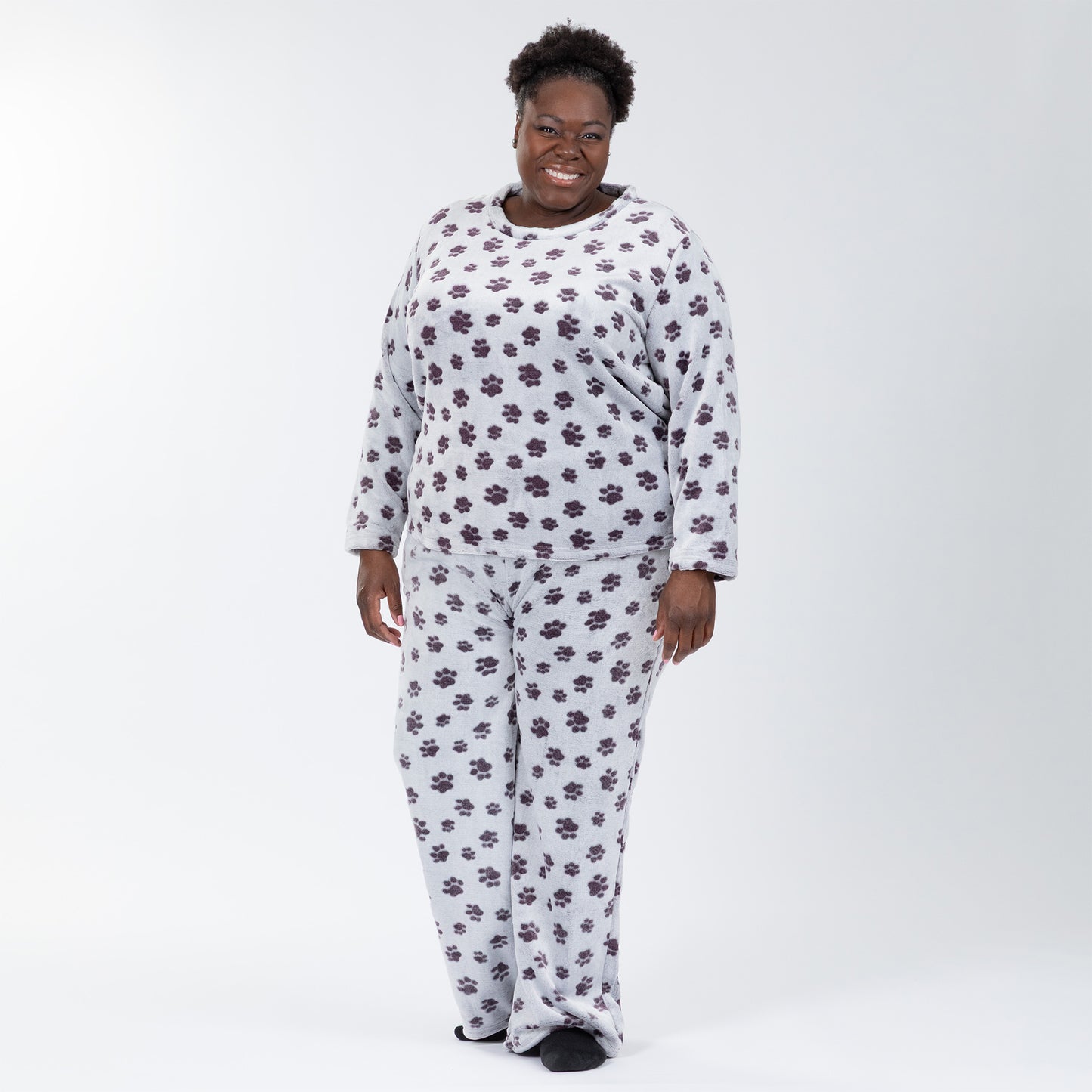 A woman wearing a matching set of fluffy, soft long-sleeve pajamas. The top and pants are light grey and patterned with purple paw prints. Plus and extended size available.