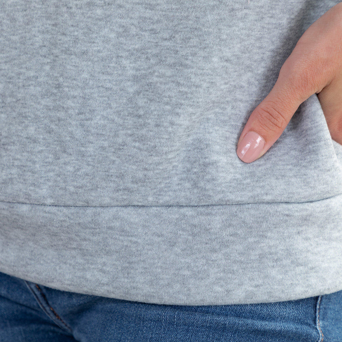 Close-up of a person wearing a heather gray sweatshirt with a side pocket, showing a hand in the pocket and blue jeans below.