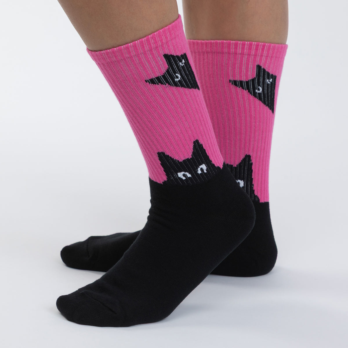 A pair of crew socks worn on a person's legs, featuring a bright pink upper with black cat silhouettes and white eyes, and a solid black lower section.