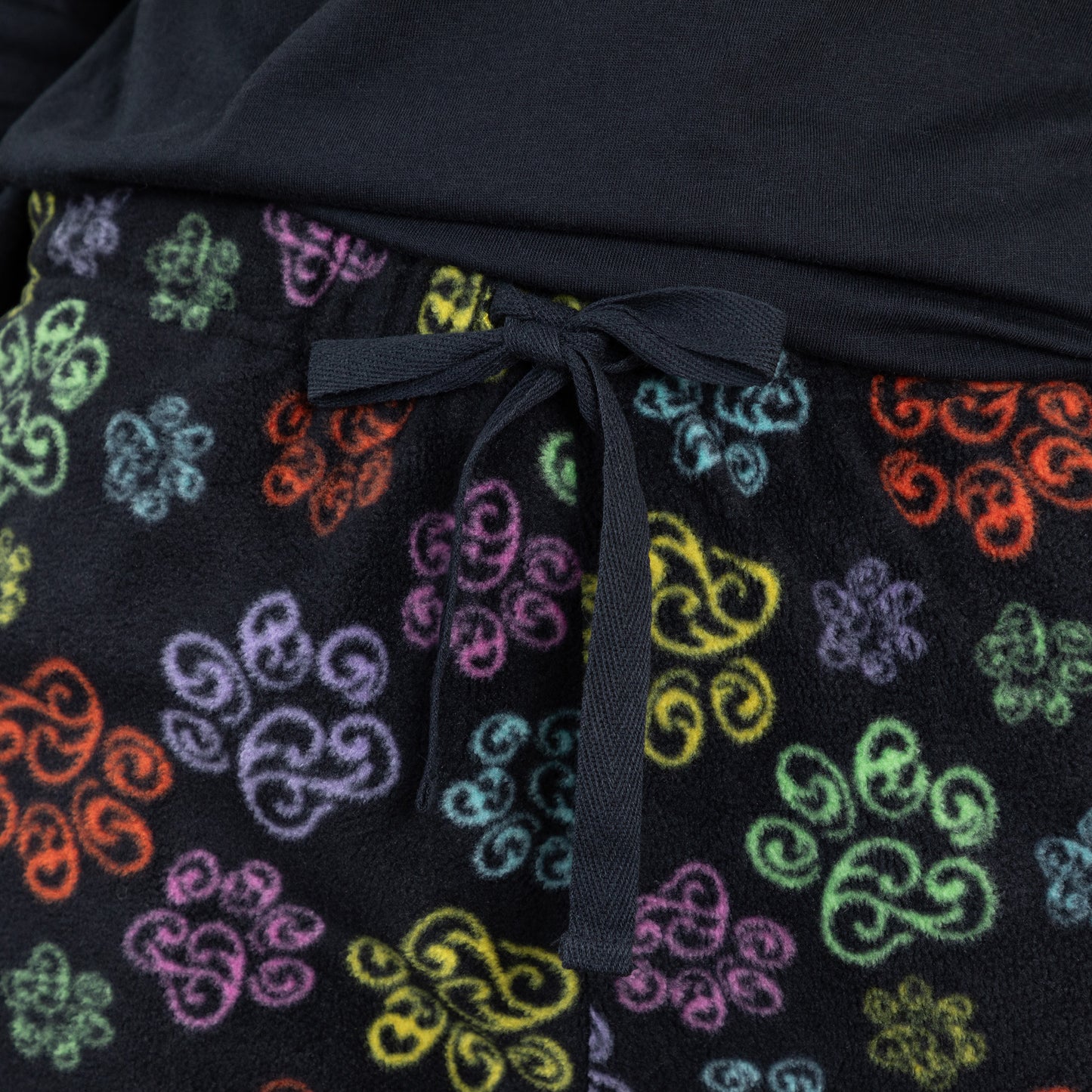 A woman standing in a bedroom setting wearing a black long-sleeve top with swirling multicolored paw print and with design swirling multicolored paw print pants showing a detail image of the adjustable drawstring pants