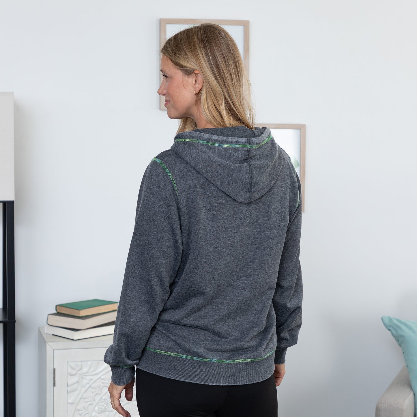 A woman shown from the back wearing a dark heather gray hoodie with thin green contrast stitching, standing in a brightly lit room.