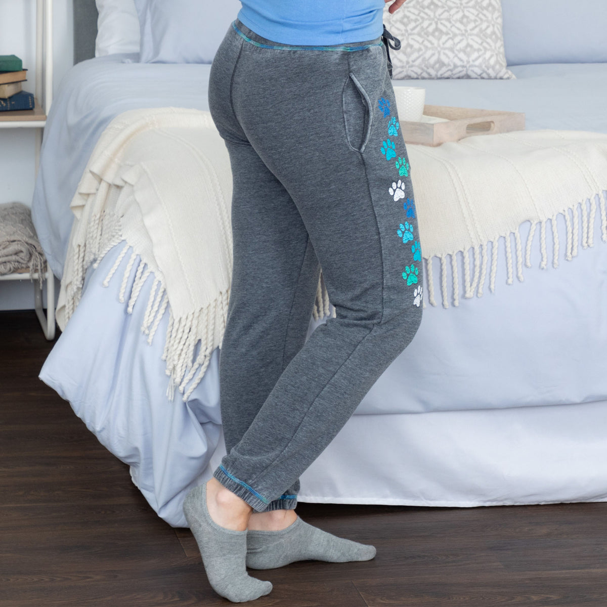 Person wearing gray joggers with colorful paw prints, standing next to a bed.