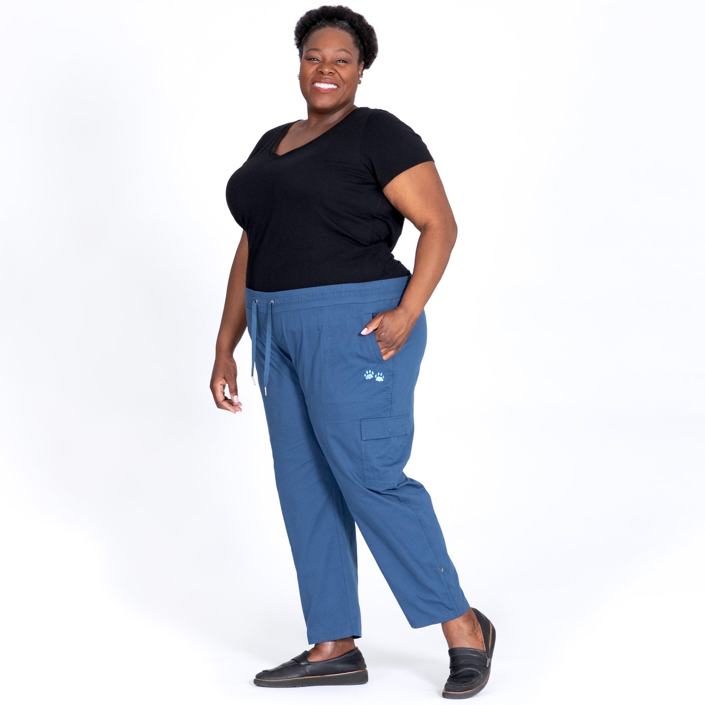 Smiling woman wearing a black shirt and blue pants, standing against a white background.