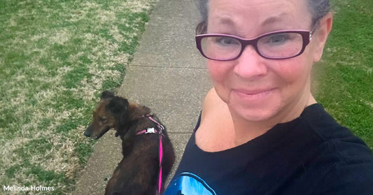 A woman wearing glasses takes a selfie while walking a brown dog on a pink leash along a suburban sidewalk at dusk.