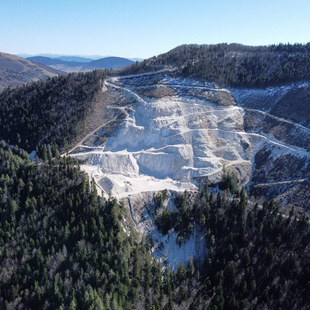 An aerial view of a large open-pit quarry carved into a forested mountainside, with snow-capped peaks visible in the distance.