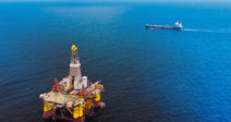 Offshore drilling platform stands in deep blue ocean with a ship visible on the horizon behind it.