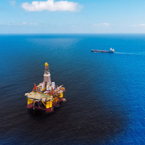Offshore drilling platform stands in deep blue ocean with a ship visible on the horizon behind it.