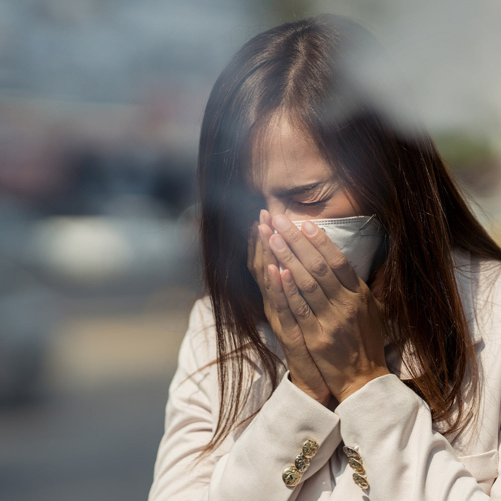 Woman wearing a face mask outdoors, covering her nose and mouth with both hands as if reacting to poor air quality.
