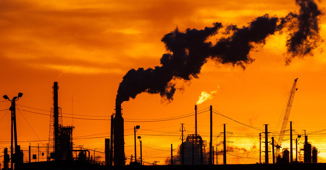 Silhouetted industrial refinery structures and smokestacks release dark smoke against a vivid orange sunset sky.
