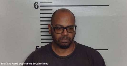 Mugshot of a man wearing glasses in front of a height chart, facing forward with a neutral expression.