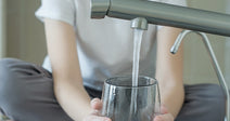 Child sits at a kitchen sink filling a clear glass with water from a modern faucet.