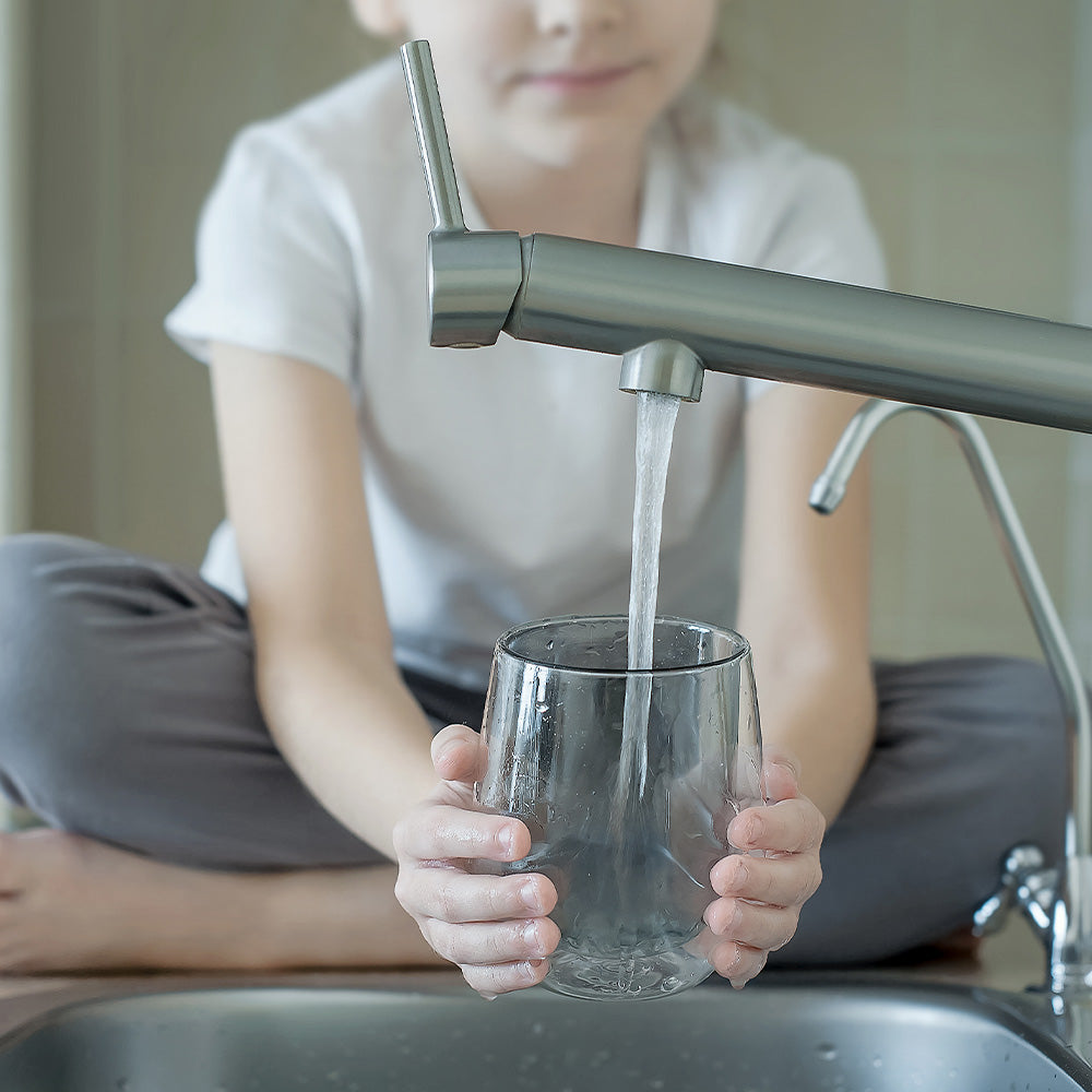 Child sits at a kitchen sink filling a clear glass with water from a modern faucet.