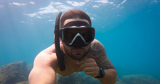 Close-up of a snorkeler underwater with mask and snorkel, arm extended toward the camera, rocky ocean floor visible below.