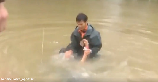 A man stands in deep, muddy floodwater while holding onto another person to help them stay above the water.