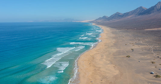 Turquoise water laps against a wide, sandy beach backed by rugged brown mountains and arid terrain stretching inland.