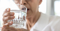 An elderly woman with white hair and glasses holds a glass of water up to her lips near a bright window.