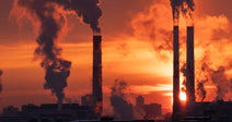 Several industrial smokestacks billow thick plumes of smoke against a vivid orange and red sunset sky over a city skyline.