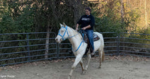 Woman riding a light-colored horse inside a fenced outdoor arena surrounded by trees.