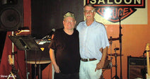 Two older men standing arm in arm on a small stage with guitars and drums behind them, beneath a “Double Saloon” sign.