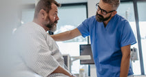 A healthcare worker in blue scrubs places a reassuring hand on a seated patient's shoulder in a modern medical examination room.