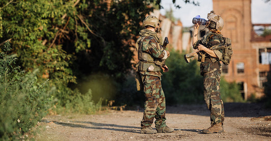 Two soldiers in camouflage uniforms and tactical gear stand outdoors, one holding a rifle while the other drinks from a water bottle near an abandoned building.