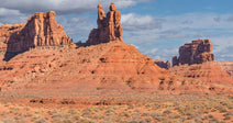 Two tall red sandstone buttes stand in a flat desert with scattered shrubs beneath a blue sky.