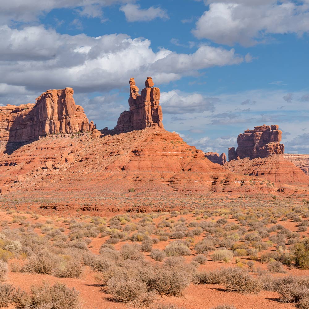 Two tall red sandstone buttes stand in a flat desert with scattered shrubs beneath a blue sky.