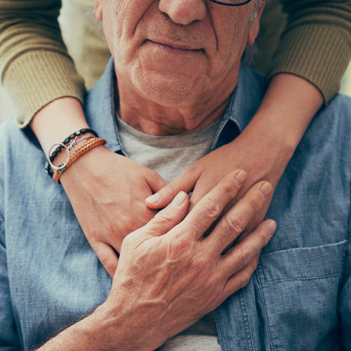 An elderly man sitting indoors is warmly embraced from behind by a younger person resting their arms across his chest.