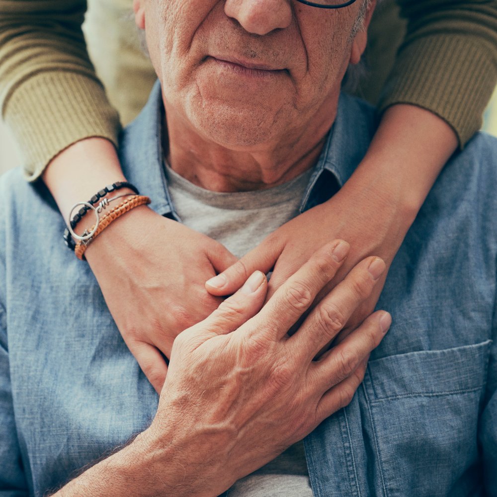 An elderly man sitting indoors is warmly embraced from behind by a younger person resting their arms across his chest.