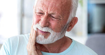 An elderly man with white hair and beard holds his jaw in pain while sitting indoors.