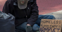 Person sitting on a sidewalk with a sign reading “Homeless Humiliated Army Vet Please Help.”