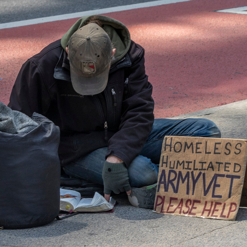 Person sitting on a sidewalk with a sign reading “Homeless Humiliated Army Vet Please Help.”