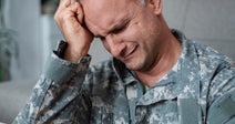 Man in military fatigues sitting with his eyes closed and his fist pressed against his forehead.