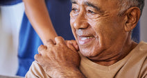 Close-up of an older man smiling while a caregiver in scrubs supports him with a hand on his shoulder.