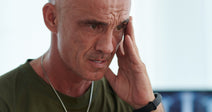 A middle-aged man wearing a military dog tag and olive green shirt holds his hand to his temple with a distressed expression, seated in a bright medical office.