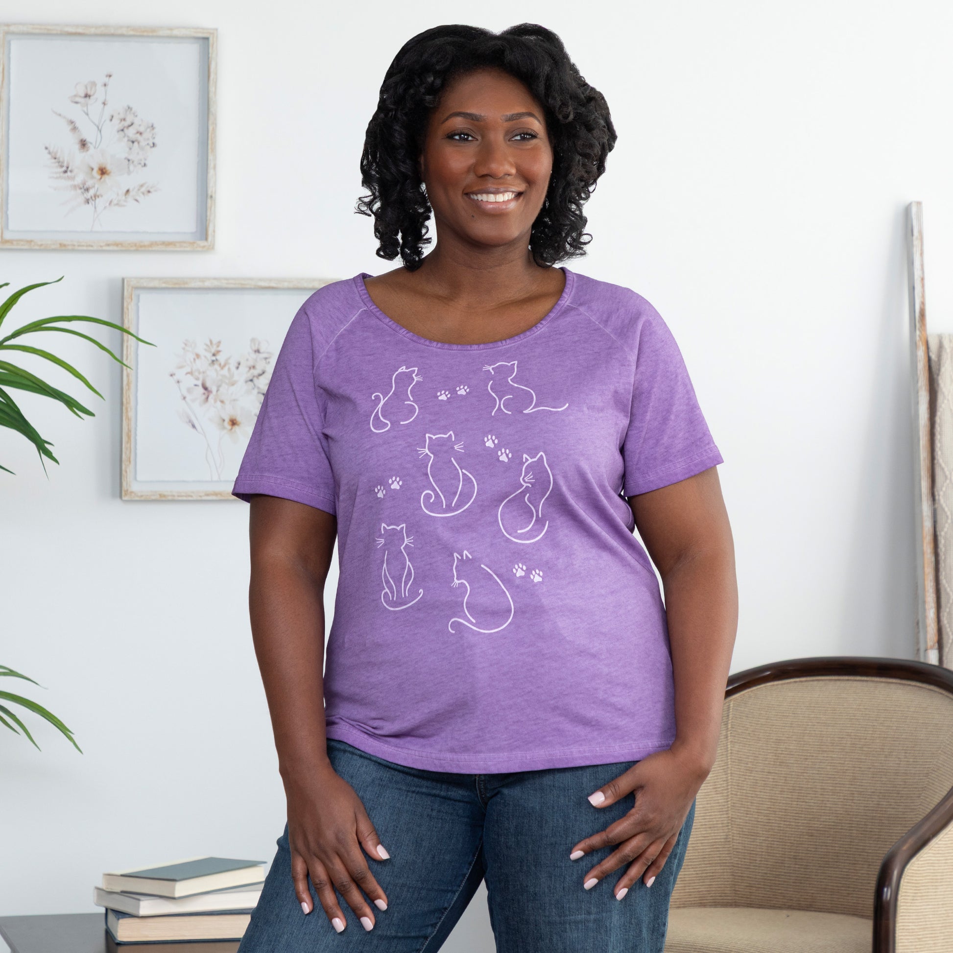 Woman wearing a purple acid washed scoop neck t-shirt with cat designs and paw prints, in a home setting.