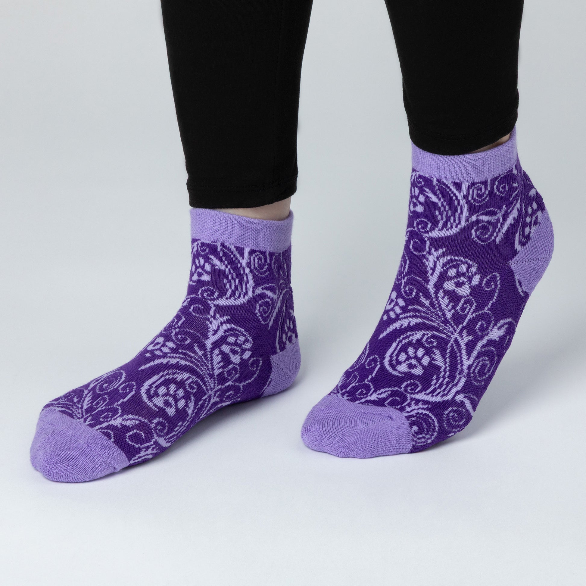 A person wearing ankle length comfortable cotton-blend socks that are dark purple in color contrasted with a lavender paw print and swirl design. The cuff, heels and toes of the sock are a plain lavender shade. 