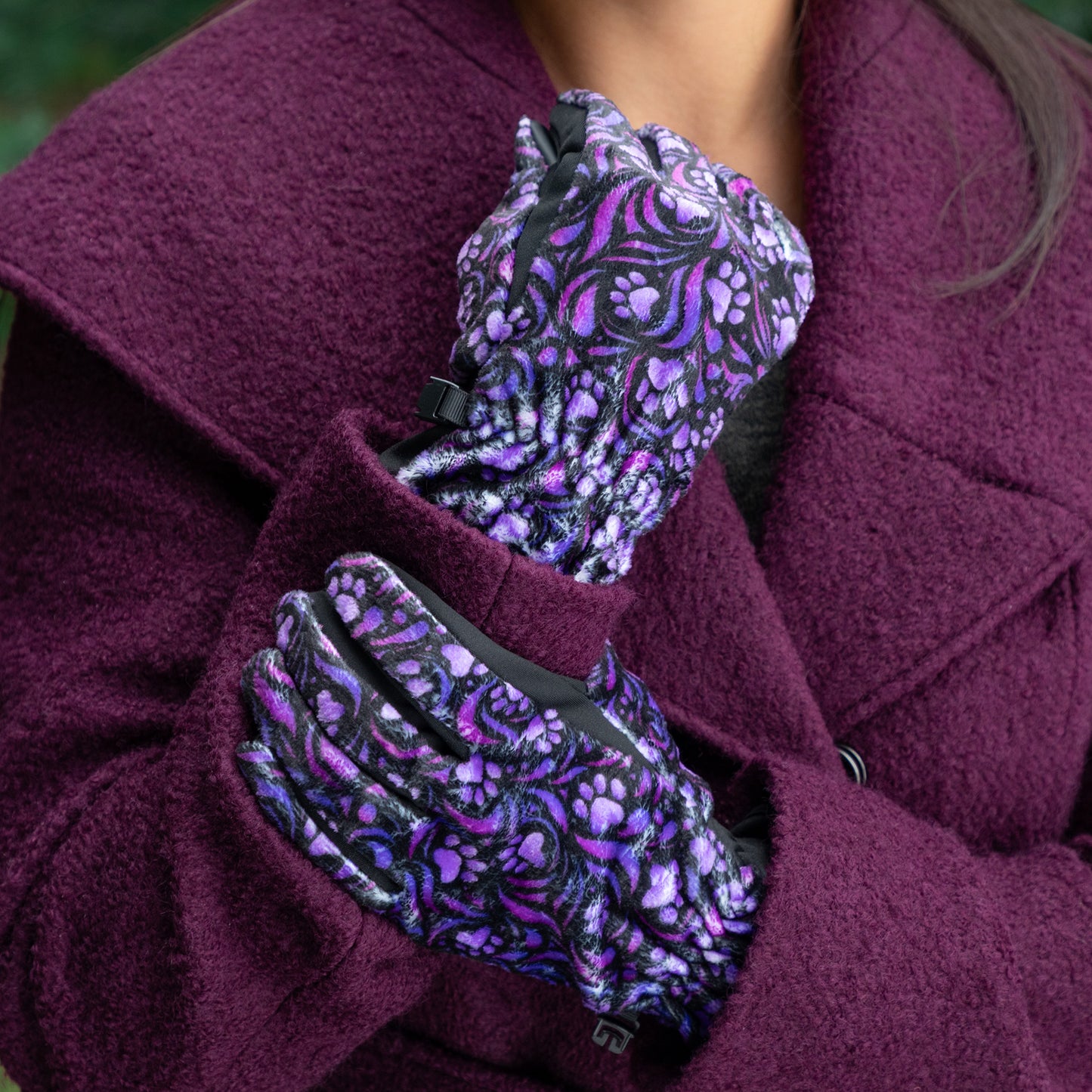 A woman, outside, wearing a coat and a pair of touchscreen technology fingertips warm black fleece gloves adorned with purple paw prints and swirls. 