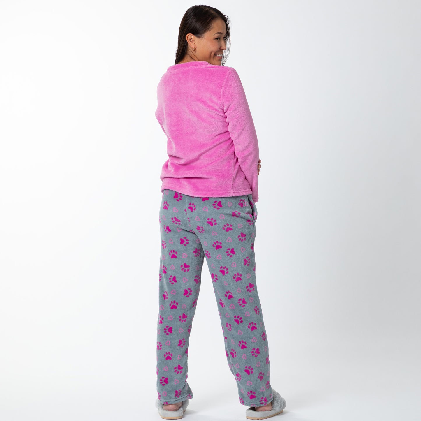 A woman wearing a pink long-sleeve top with an embroidered dog face design and gray pants with pink paw prints and hearts pattern. She is standing against white background showing the back of the set. Design a pink dog in super cozy fabric, showing an Elastic waistband with a drawstring, two side pockets 