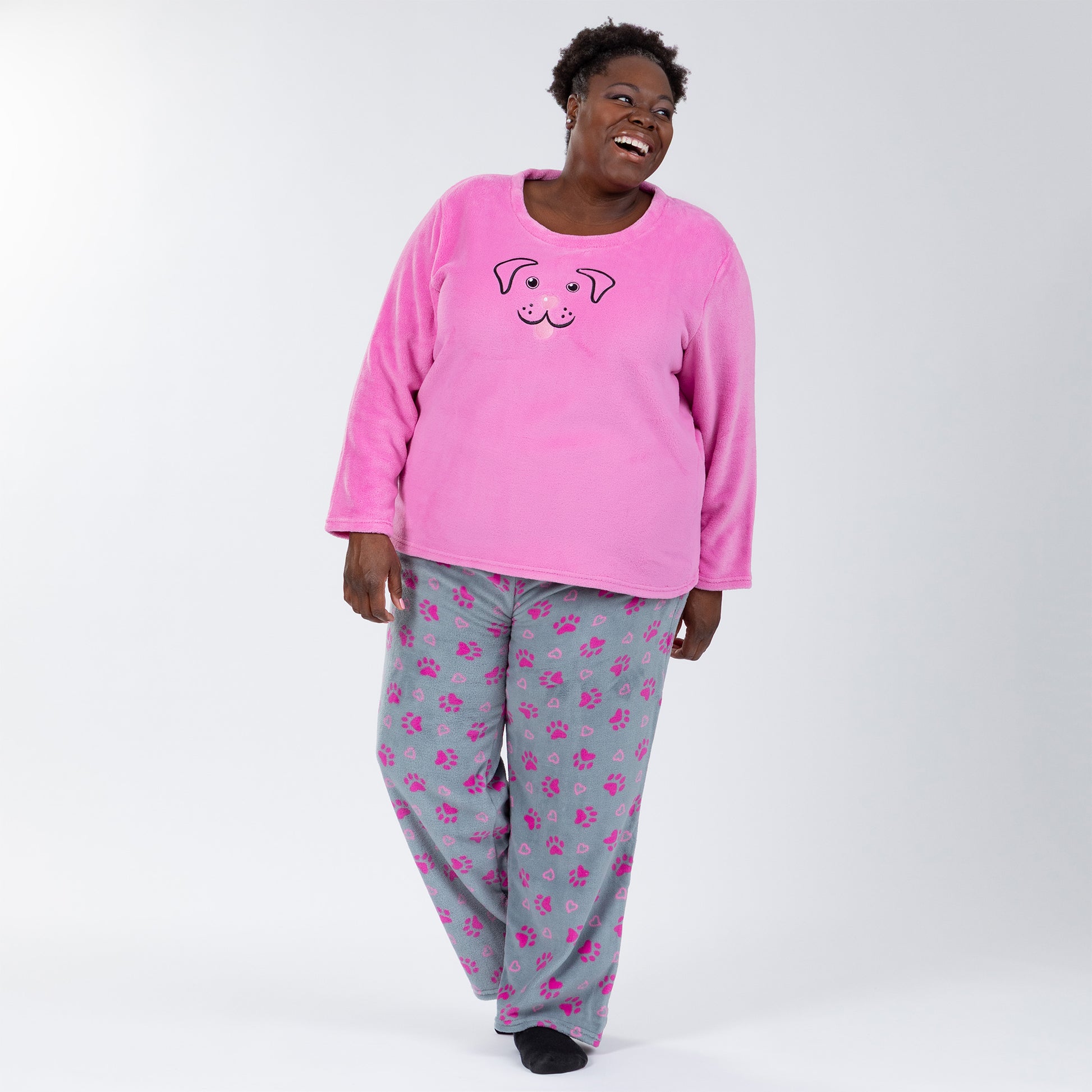 A woman wearing a pink long-sleeve top with an embroidered dog face design and gray pants with pink paw prints and hearts pattern. She is standing against white background showing the back of the set. Design a pink dog in super cozy fabric, showing an Elastic waistband with a drawstring, two side pockets . Extended and plus size available. 