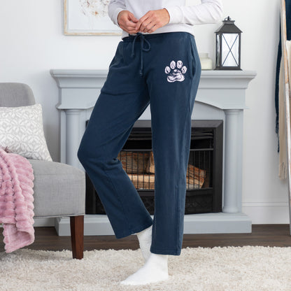 A person wearing navy blue lounge pants with a white paw print graphic on the upper left leg, standing in a living room in front of a fireplace.