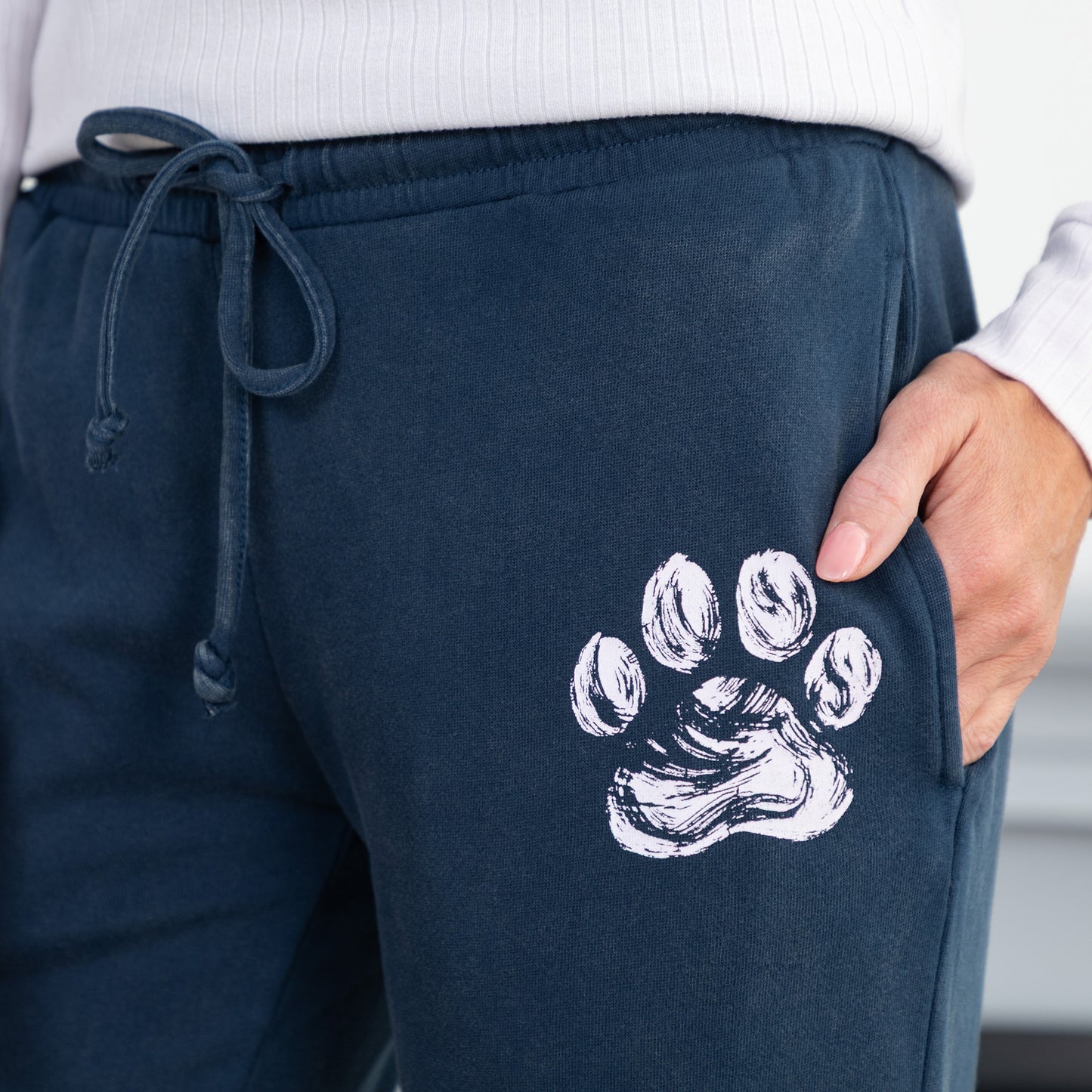 Close-up of a person wearing navy blue sweatpants with a white paw print graphic on the thigh, paired with a white ribbed top. The person's hand is resting in the pocket.