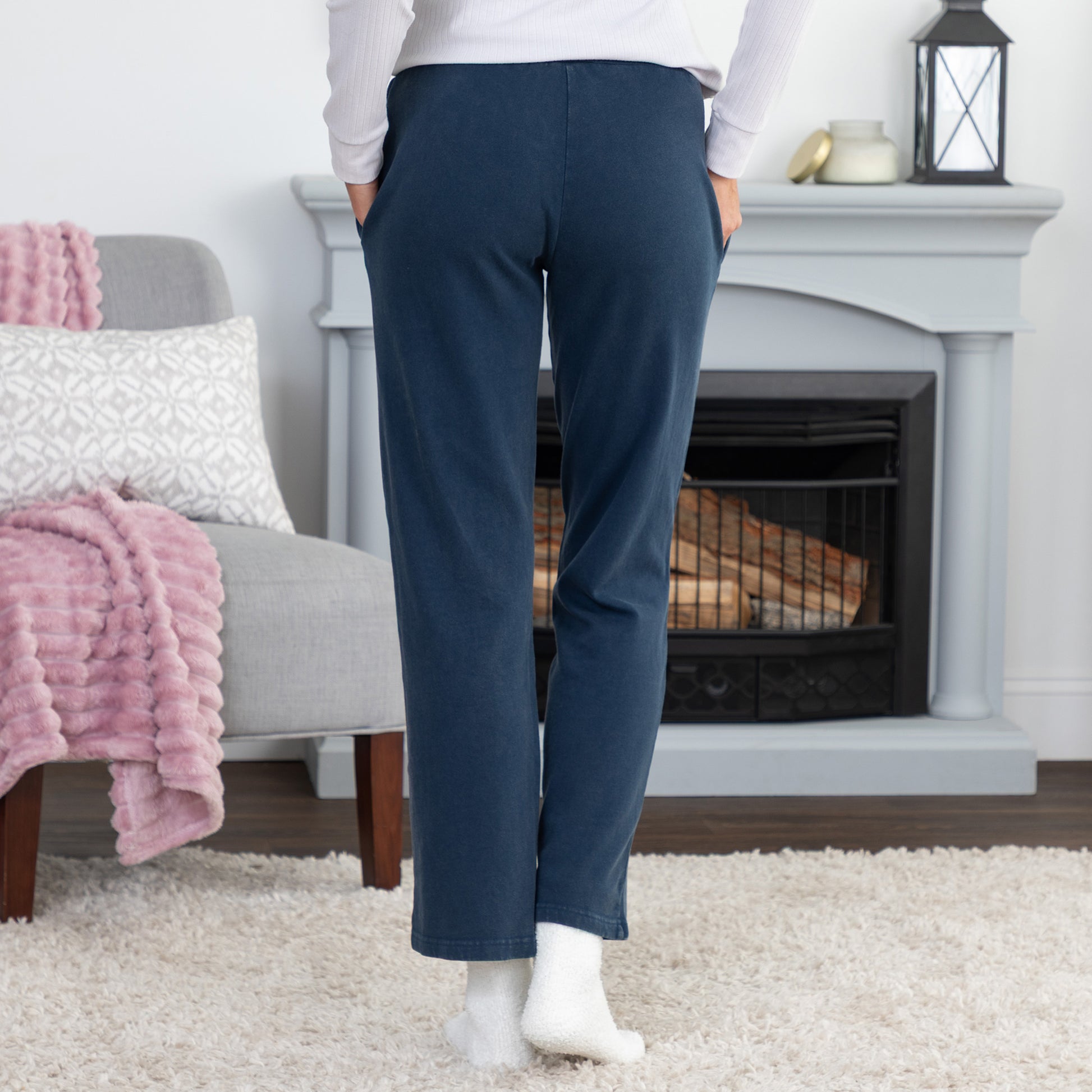 A person standing in a living room wearing navy blue straight-leg pants, a white long-sleeve top, and white socks, with a fireplace and cozy chair in the background.