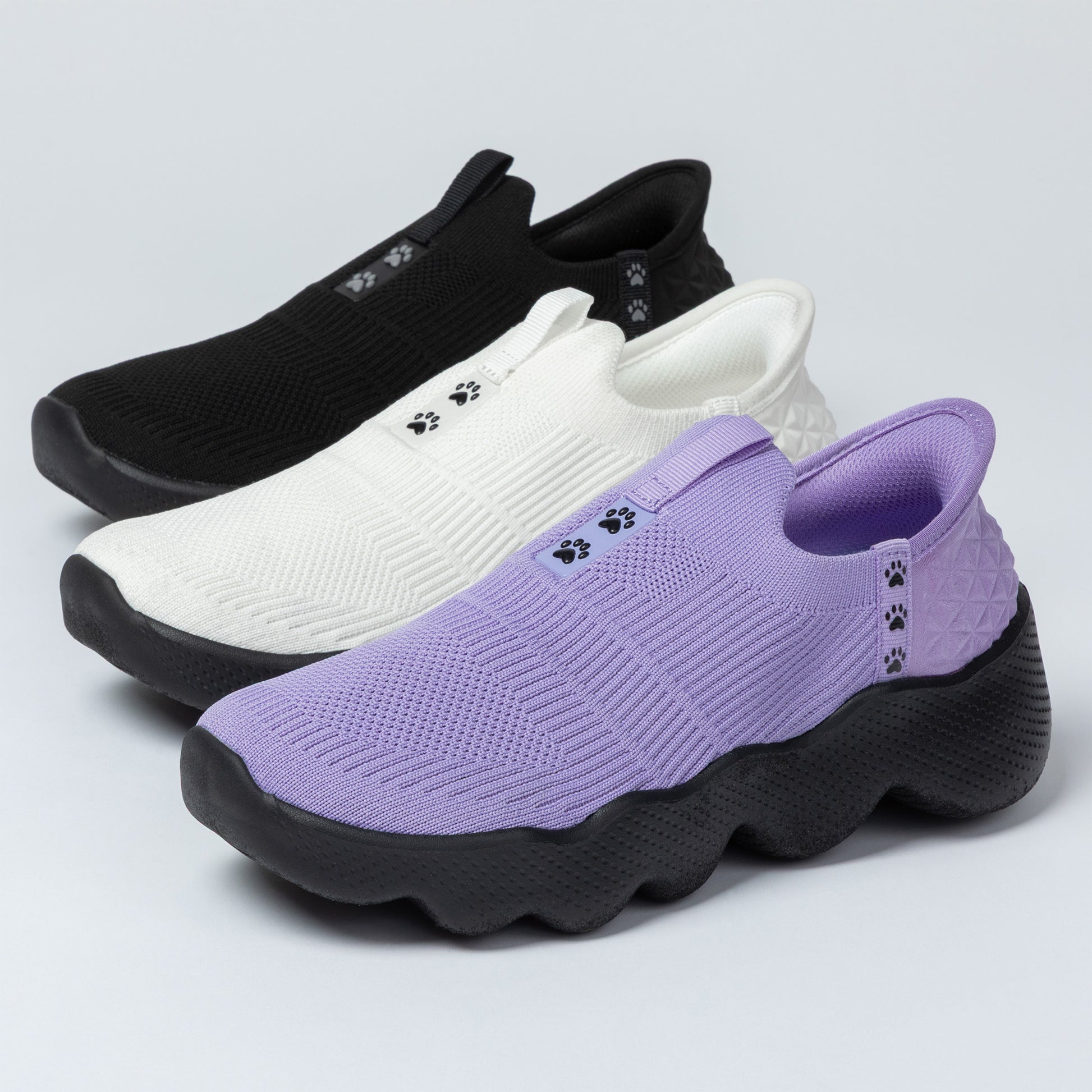Purple slip-on shoes with paw prints, black and white shoes in the background on a light gray background