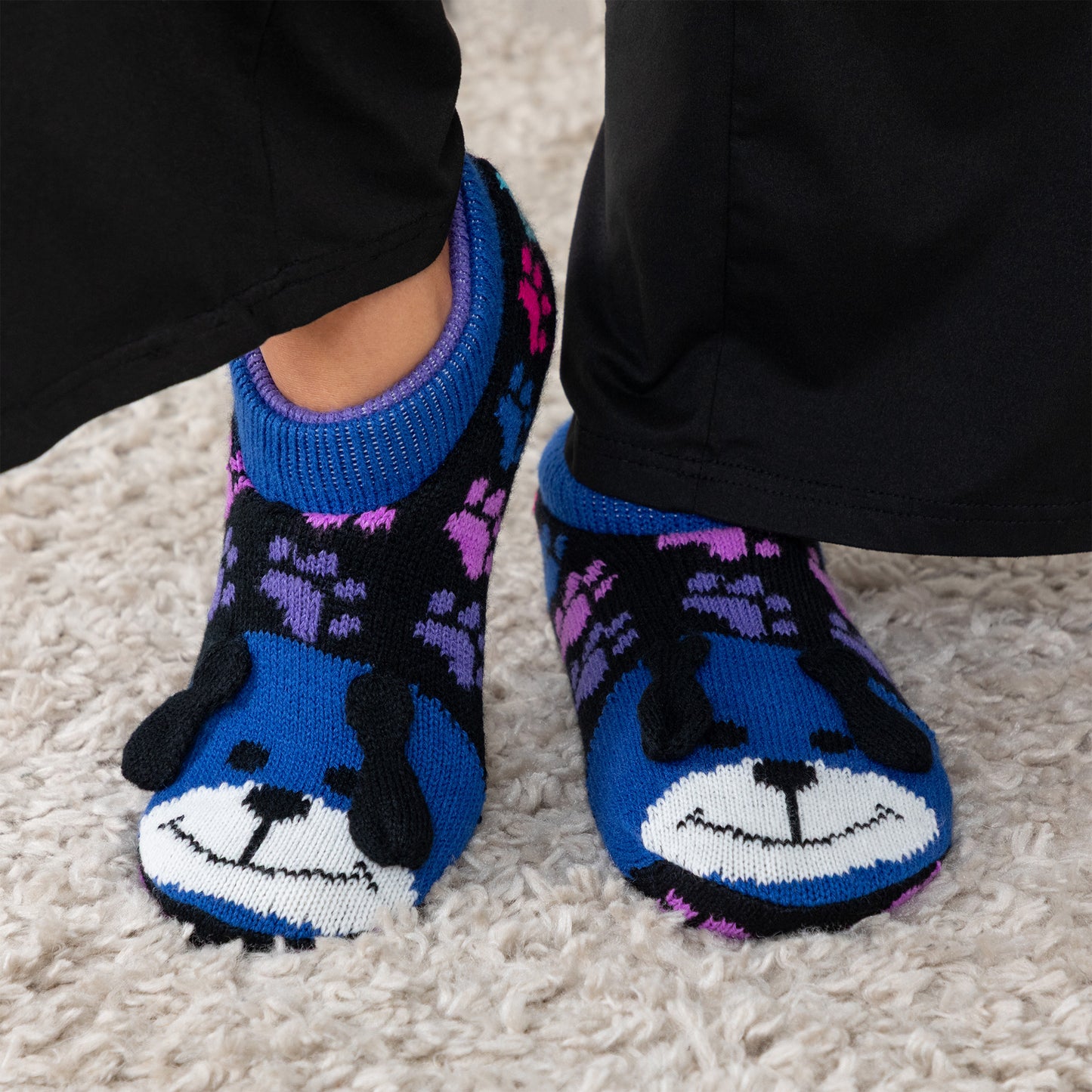 Person wearing blue and rainbow paw patterned knitted slipper socks with a smiling dog face on top, on a carpet floor rainbow dog