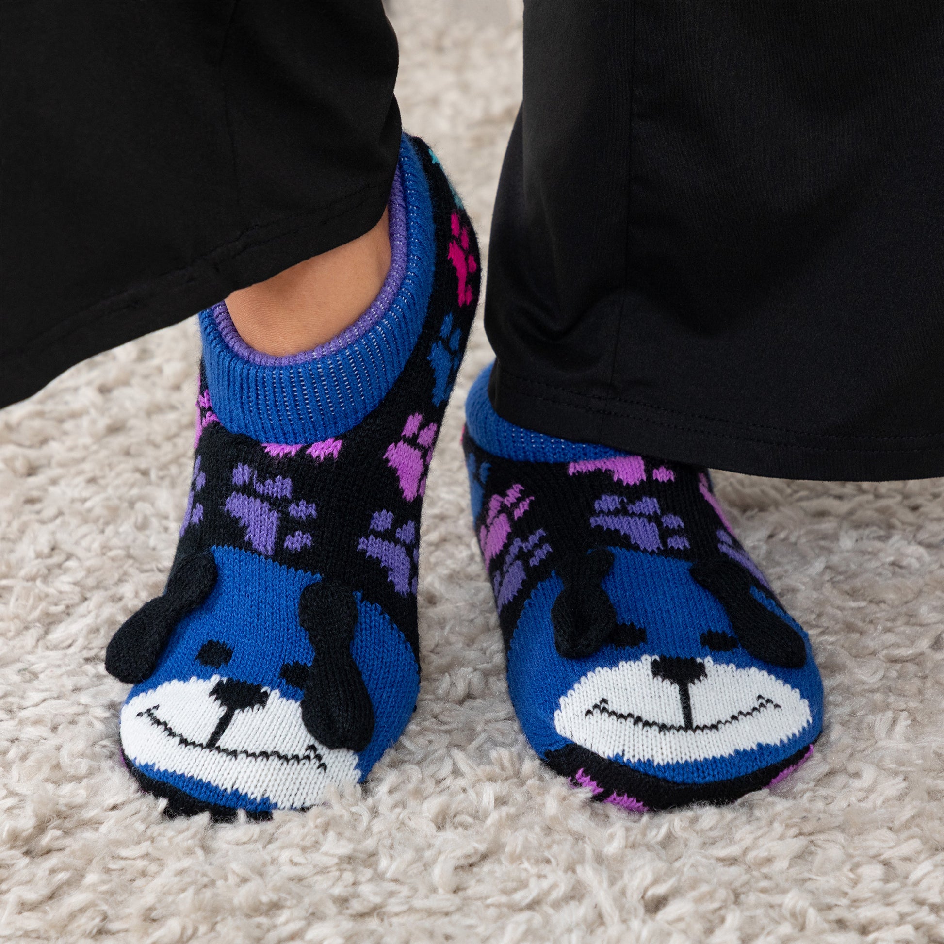 Person wearing blue and rainbow paw patterned knitted slipper socks with a smiling dog face on top, on a carpet floor rainbow dog