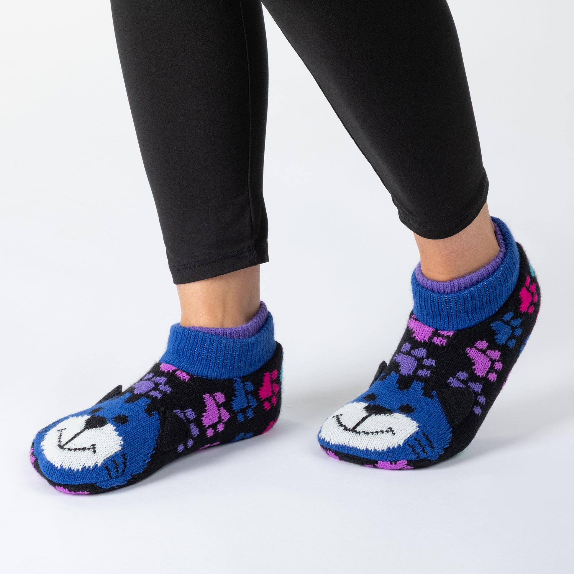 A person wearing black leggings and blue knit booties with a cat face design and purple pattern details, against a white background.