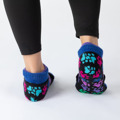 A person wearing black leggings and colorful knit slip-on socks with a blue cuff, shown from the back against a light gray studio background.