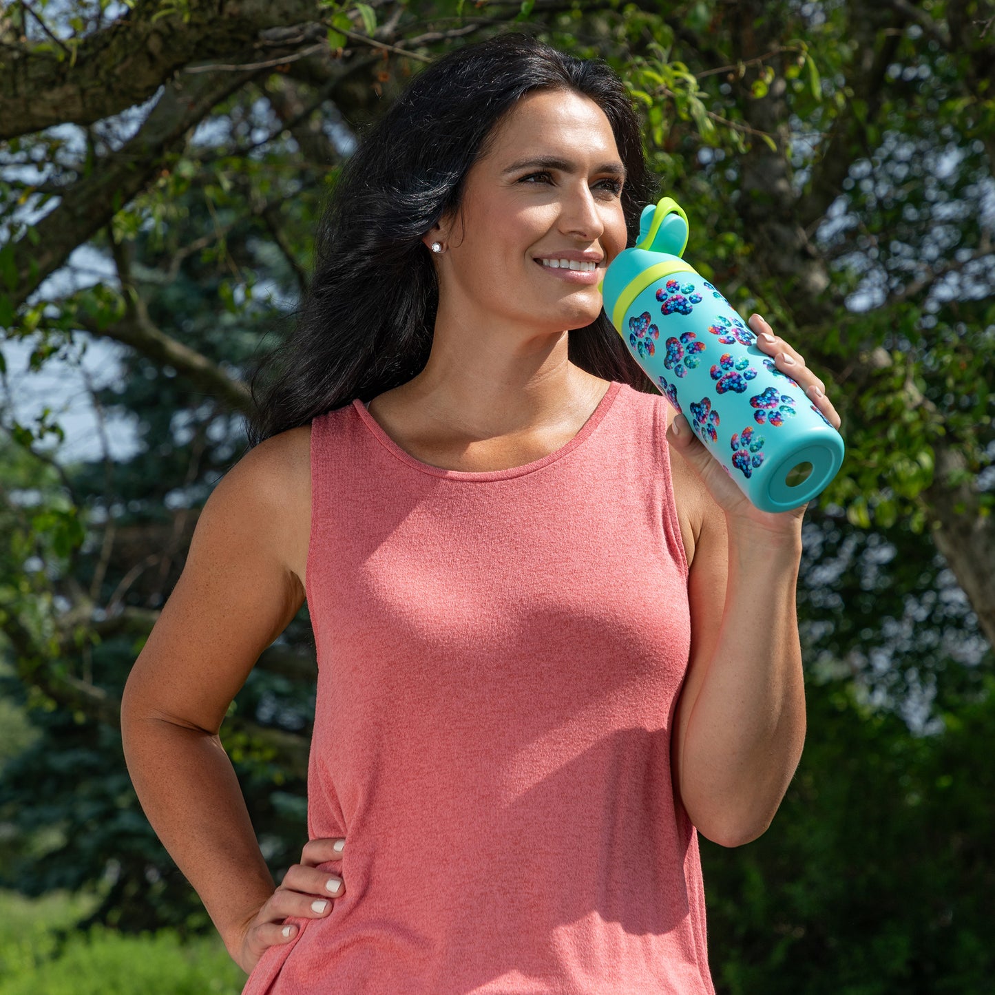 Woman in a pink tank top holds a colorful water bottle outdoors, smiling confidently.